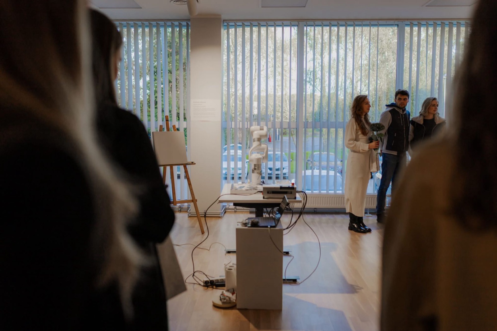 Exhibition view at Imanta Culture Centre with the robotic arm, an easel and visitors by the window